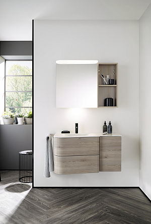Ein Badezimmer, in dem der Burgbad Badu Waschtisch in Holzoptik an einer weißen Wand montiert ist, begleitet von einem beleuchteten Spiegel.