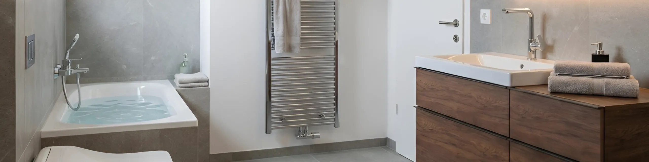 Ein harmonisches Badezimmer mit eingelassener Badewanne, Handbrause und modernem Holz-Waschtisch. Ein verchromter Handtuchheizkörper hängt an der Wand, daneben liegt ein gefaltetes Handtuch auf der Ablage. Die Gestaltung wirkt warm und hochwertig.