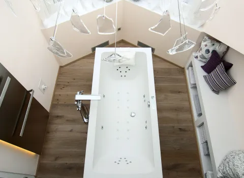 Eine rechteckige Whirlwanne, umgeben von edlem Holzfußboden und sanfter Beleuchtung, dominiert das Badezimmer. Glaselemente hängen darüber, und Sitzbänke mit Kissen verleihen dem Raum Gemütlichkeit.