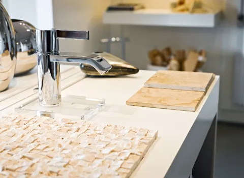 Ein glänzender, moderner Wasserhahn aus Chrom steht auf einer weißen Oberfläche neben sandfarbenen Natursteinfliesen und einer strukturierten Steinplatte.