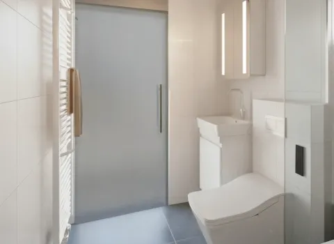 Ein minimalistisches Badezimmer mit schmaler Raumaufteilung, weißem Waschtisch, wandhängendem WC und Spiegelschrank mit LED. Der seitlich montierte Heizkörper trägt ein beigefarbenes Handtuch.