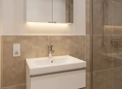 Ein moderner Waschtisch mit zwei Schubladen steht vor einer Wand mit beigen Fliesen, darüber ein beleuchteter Spiegelschrank mit integrierter LED-Leiste. Rechts daneben befindet sich eine Dusche mit Glasabtrennung und Regenduschkopf. Die Kombination aus warmen Naturtönen und minimalistischem Design wirkt harmonisch und hochwertig. Eine Steckdose neben dem Waschbecken bietet zusätzlichen Komfort.