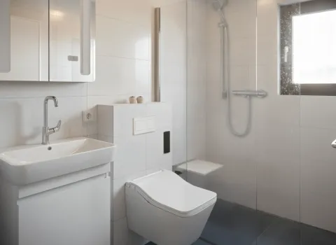 Ein kleines, barrierefreies Badezimmer mit wandhängendem WC, Glasdusche und klappbarem Duschsitz. Der Waschplatz mit Spiegelschrank ist platzsparend integriert. Helle Fliesen und ein Fenster sorgen für Licht und Weite.