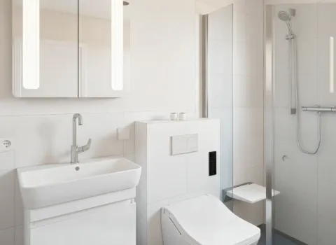 Das Bild zeigt ein helles Bad mit kompaktem Waschbecken, wandmontiertem WC und bodengleicher Dusche mit Klappsitz. Der Spiegel mit LED-Beleuchtung ergänzt das moderne Design.