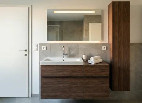 Moderner Waschtisch mit Unterschrank und Hochschrank in Holzoptik, integriertem Spiegel mit Beleuchtung und grauen Wandfliesen.