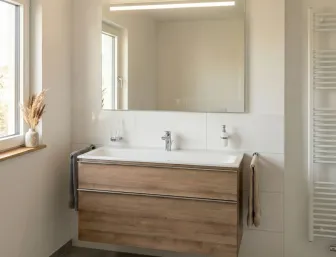 Grosser Waschtisch mit Holzschubladen in einem minimalistischen Badezimmer. Der grosse Leuchtspiegel darüber reflektiert Teile des Raums.