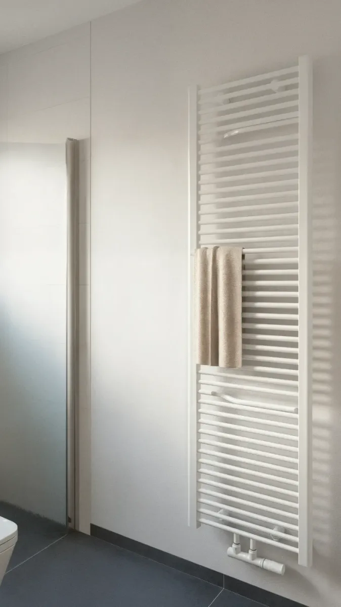 Ein vertikaler Heizkörper im Badezimmer dient gleichzeitig als Handtuchhalter. Das Design ist schlicht und funktional, perfekt für kleine oder moderne Bäder. Links ist eine Dusche mit Glastür angeschnitten sichtbar.