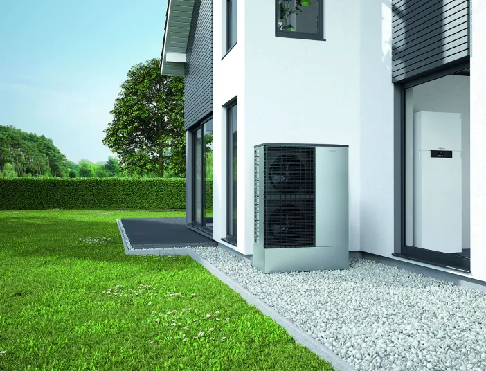 Eine kompakte Wärmepumpen-Außeneinheit ist neben einem Neubau installiert und ruht auf einem Metallständer über einem Kiesbett.